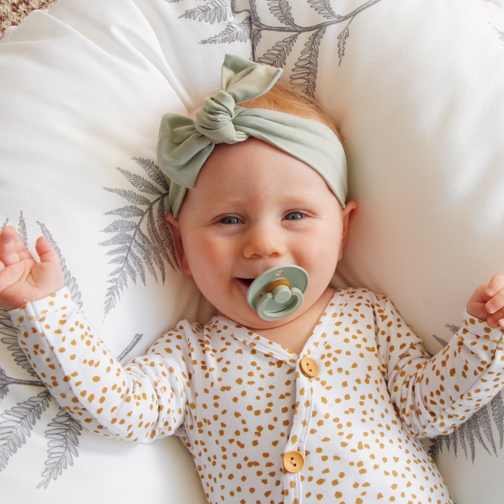 A baby wearing a sage green bow knot headband with a white patterned blanket in the background.