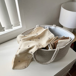 Folded beige blanket in a woven basket on a white surface with a lamp and window in the background.